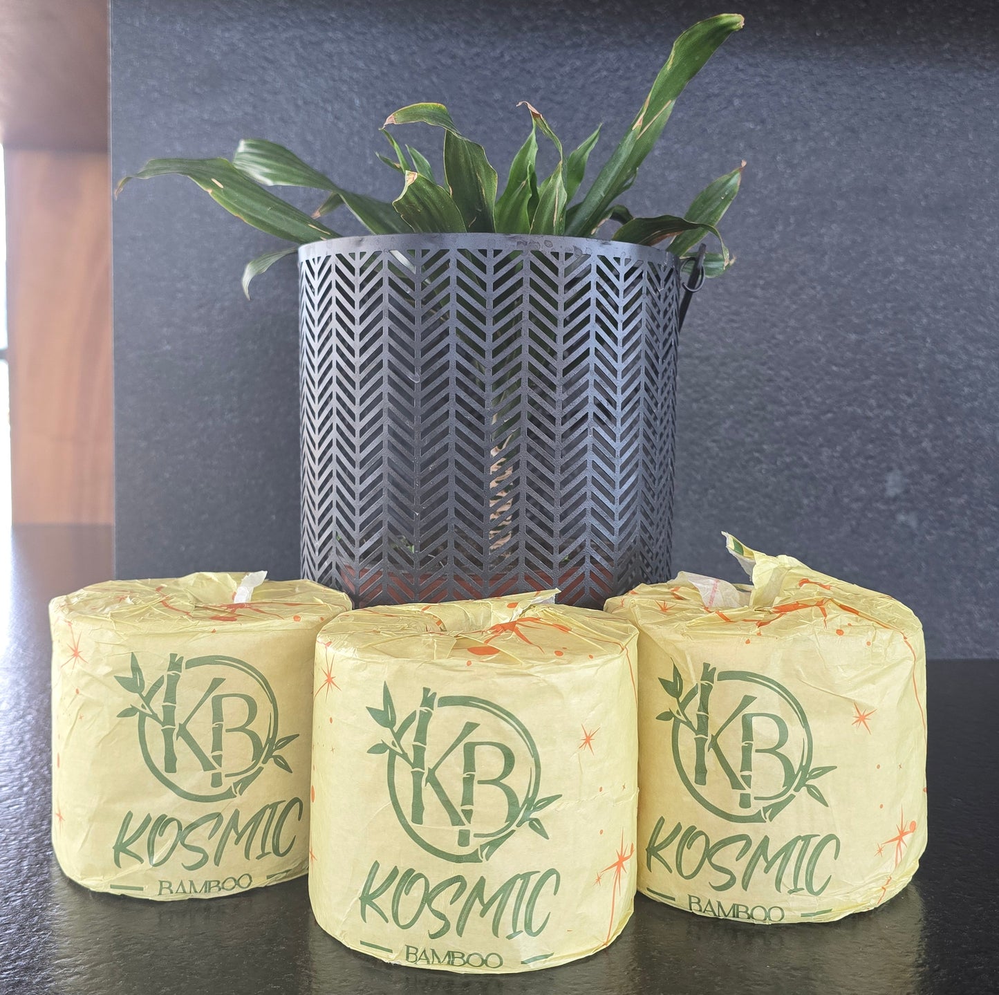Three kosmic bamboo bathroom tissue rolls on a dark surface with a plant in the baackground